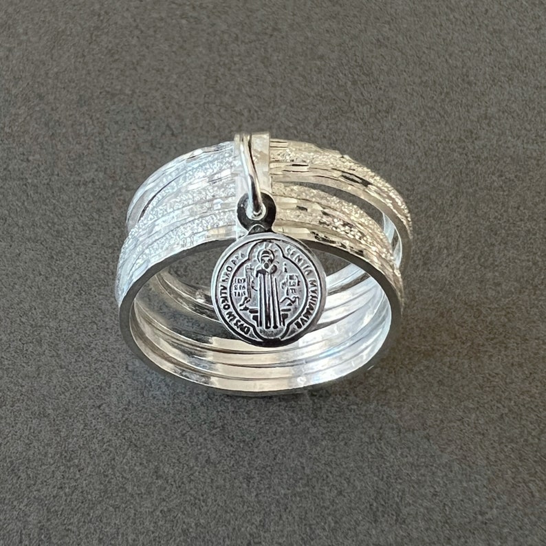 May include: Silver ring with a Saint Benedict medal charm. The ring is made of multiple bands with a textured finish.