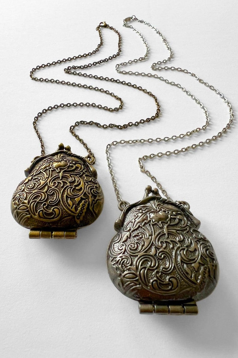 Puede incluir: Dos colgantes ornamentados, de color dorado, con bisagras, de estilo medall&oacute;n, cuelgan de una cadena plateada. Los colgantes tienen forma de monedero y presentan intrincados dise&ntilde;os florales.