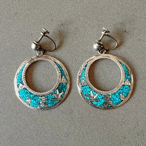 May include: Pair of silver-toned hoop earrings with turquoise inlay. The earrings feature a circular design with intricate details and a screw-back closure. The turquoise stones are set within a silver frame, creating a striking contrast. The earrings are a vintage style.