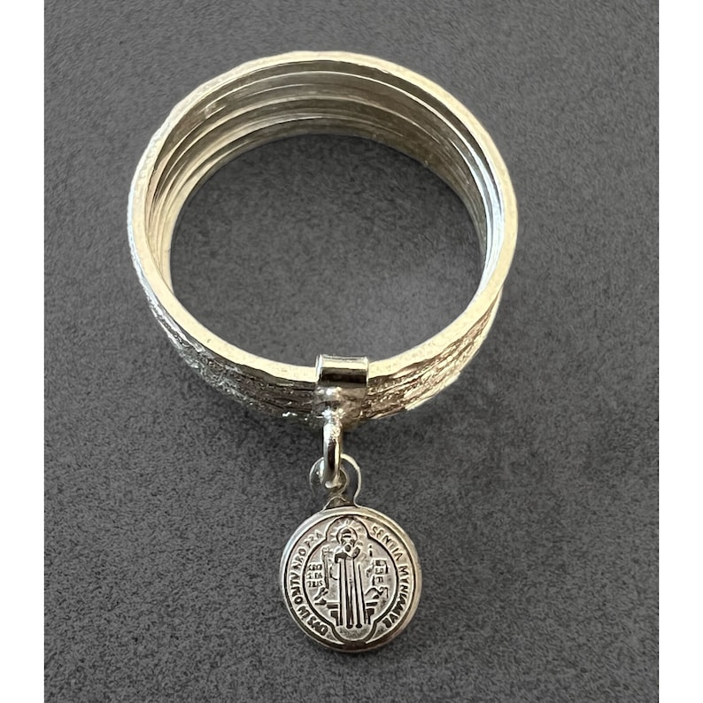 May include: A silver ring with a Saint Christopher medal charm. The ring is made of multiple bands of silver, giving it a textured look.