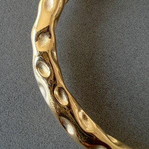 May include: A gold-toned metal bracelet with a textured, hammered finish. The bracelet is curved and has a smooth, polished surface.