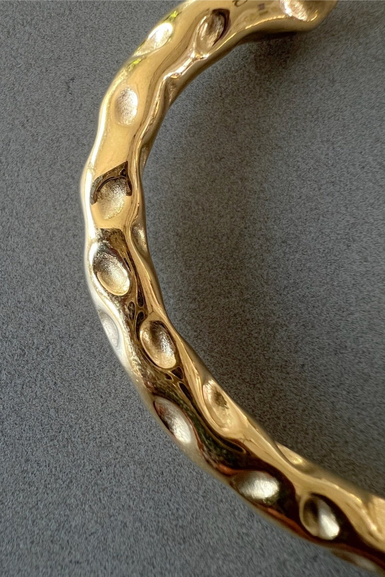 May include: A gold-toned metal bracelet with a textured, hammered finish. The bracelet is curved and has a smooth, polished surface.