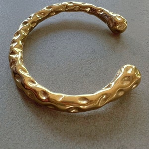 May include: A gold-toned bangle bracelet with a textured, organic design. The bracelet is open and has a slightly curved shape.