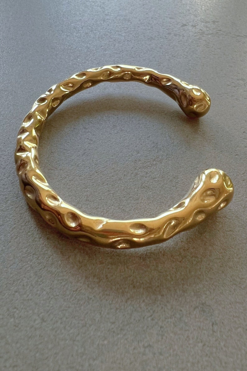 May include: A gold-toned bangle bracelet with a textured, organic design. The bracelet is open and has a slightly curved shape.
