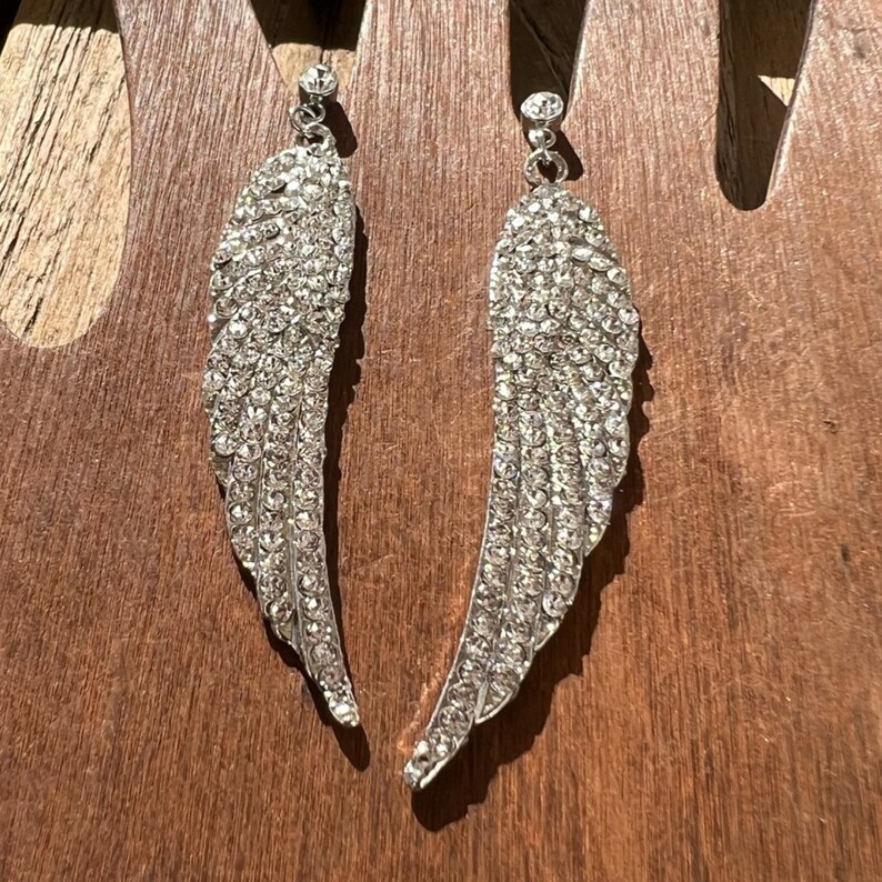May include: A pair of silver dangle earrings with crystal wings. The wings are made of small, clear crystals that are set in a silver metal.