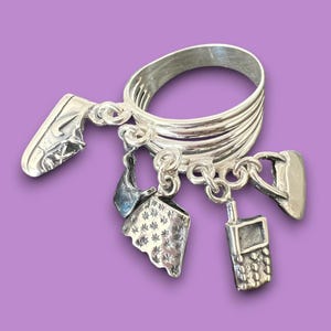 May include: A silver ring with multiple bands and five charms hanging from it. The charms are a shoe, a cell phone, a house, a star, and a heart.