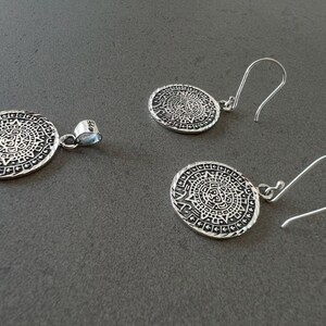 May include: A silver pendant and matching earrings set. The jewelry features an oval design with an intricate pattern, possibly inspired by ancient art. The earrings have a hook closure. The set is displayed on a gray surface.
