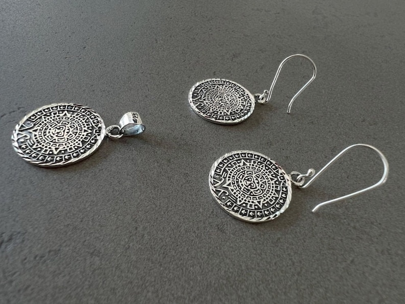May include: A silver pendant and matching earrings set. The jewelry features an oval design with an intricate pattern, possibly inspired by ancient art. The earrings have a hook closure. The set is displayed on a gray surface.