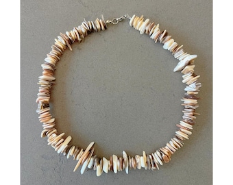 Puede incluir: Un collar hecho de conchas naturales en tonos marrones, blancos y beige. Las conchas están dispuestas en un patrón escalonado, creando un aspecto único y texturizado.