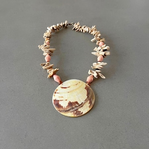 Op de afbeelding: Een ketting met een grote, ronde schelphanger in crème en bruin. Het snoer bestaat uit onregelmatige, lichtgekleurde schelpfragmenten afgewisseld met gladde, ovale, lichtroze kralen, afgesloten met een metalen sluiting.