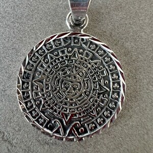 May include: A silver pendant with a detailed Mayan calendar design. The round pendant features intricate carvings and a loop for a chain. The pendant is marked with "925" on the bail, indicating its silver content. The design includes a central face.