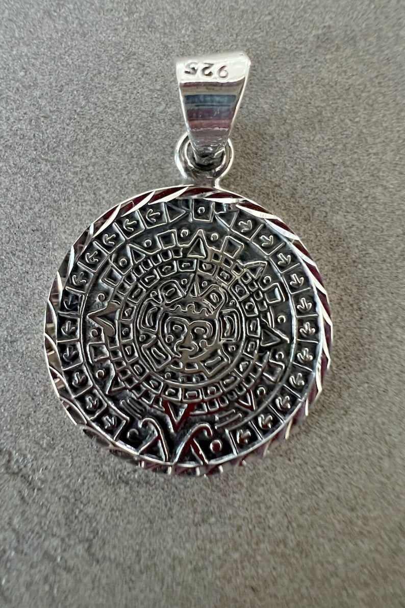 May include: A silver pendant with a detailed Mayan calendar design. The round pendant features intricate carvings and a loop for a chain. The pendant is marked with "925" on the bail, indicating its silver content. The design includes a central face.