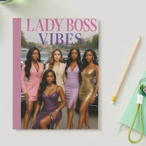 May include: A notebook with the text "LADY BOSS VIBES" in pink and purple. The cover features a group of women in business attire. A pencil, a green keychain, and a small green notebook are on the white surface.