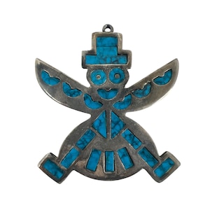 May include: A silver-toned pendant in the shape of a stylized figure with outstretched arms and legs. The figure is inlaid with turquoise-colored stones. The pendant has a loop at the top for hanging.