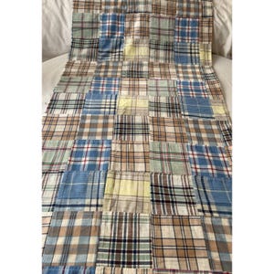 May include: A rectangular patchwork quilt featuring a grid of various plaid fabric squares in shades of blue, tan, brown, yellow, and white. The textile is composed of multiple stitched fabric patches arranged in a repeating pattern.