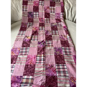 May include: A rectangular patchwork quilt featuring a grid of square fabric pieces in various shades of pink, magenta, and purple. The design alternates between solid pink squares, plaid patterns, and floral or mottled fabric prints.