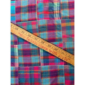 May include: A patchwork fabric featuring a multi-colored plaid pattern in shades of blue, red, purple, and orange. A wooden ruler is placed diagonally across the fabric, showing measurements from 13 to 22 inches in length.