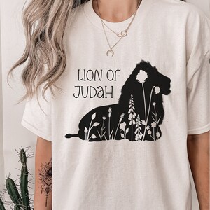 以下が含まれることがあります： 白いTシャツに、花が咲いたライオンの黒いシルエットと「Lion of Judah」の文字が前面にプリントされています。