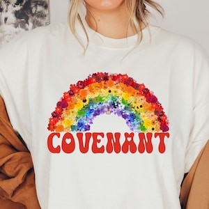 以下が含まれることがあります： 花でできた虹と、ハートが「O」に入っている赤い「COVENANT」という言葉が書かれた白いTシャツ。