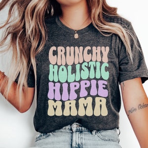 Puede incluir: Una camiseta gris oscuro con el texto "CRUNCHY HOLISTIC HIPPIE MAMA" en una fuente retro colorida.