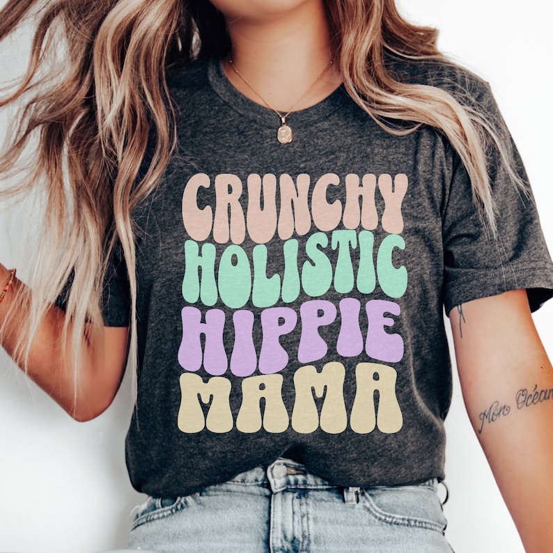 Puede incluir: Una camiseta gris oscuro con el texto "CRUNCHY HOLISTIC HIPPIE MAMA" en una fuente retro colorida.