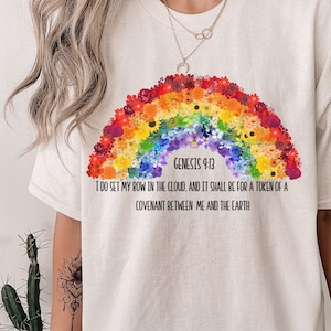 May include: Cream-colored t-shirt featuring a rainbow floral design. The rainbow is composed of various colorful flowers. Text on the shirt reads "Genesis 9:13" and a quote about a covenant.