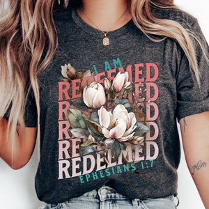 Puede incluir: Una camiseta gris con un dise&ntilde;o floral y el texto "I AM REDEEMED EPHESIANS 1:7" en rosa, rojo y azul.