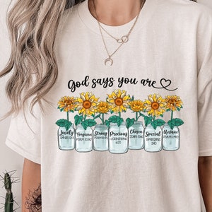 May include: A white t-shirt with the text "God says you are" and a heart symbol.  The shirt features a row of six mason jars with sunflowers growing out of them. Each jar has a different word and bible verse printed on it: "Lovely" (Daniel 12:3), "Forgiven" (Psalm 103:12), "Strong" (Psalm 18:35), "Precious" (1 Corinthians 6:20), "Chosen" (John 15:46), "Special" (Ephesians 2:10), and "Unique" (Psalm 139:13).