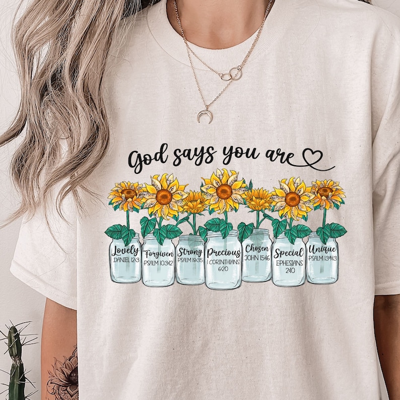 May include: A white t-shirt with the text "God says you are" and a heart symbol.  The shirt features a row of six mason jars with sunflowers growing out of them. Each jar has a different word and bible verse printed on it: "Lovely" (Daniel 12:3), "Forgiven" (Psalm 103:12), "Strong" (Psalm 18:35), "Precious" (1 Corinthians 6:20), "Chosen" (John 15:46), "Special" (Ephesians 2:10), and "Unique" (Psalm 139:13).