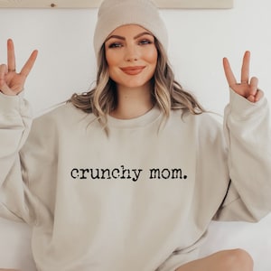 May include: A beige sweatshirt with the text "crunchy mom." printed in black on the front. The model is wearing a beige beanie and is making a peace sign with both hands.