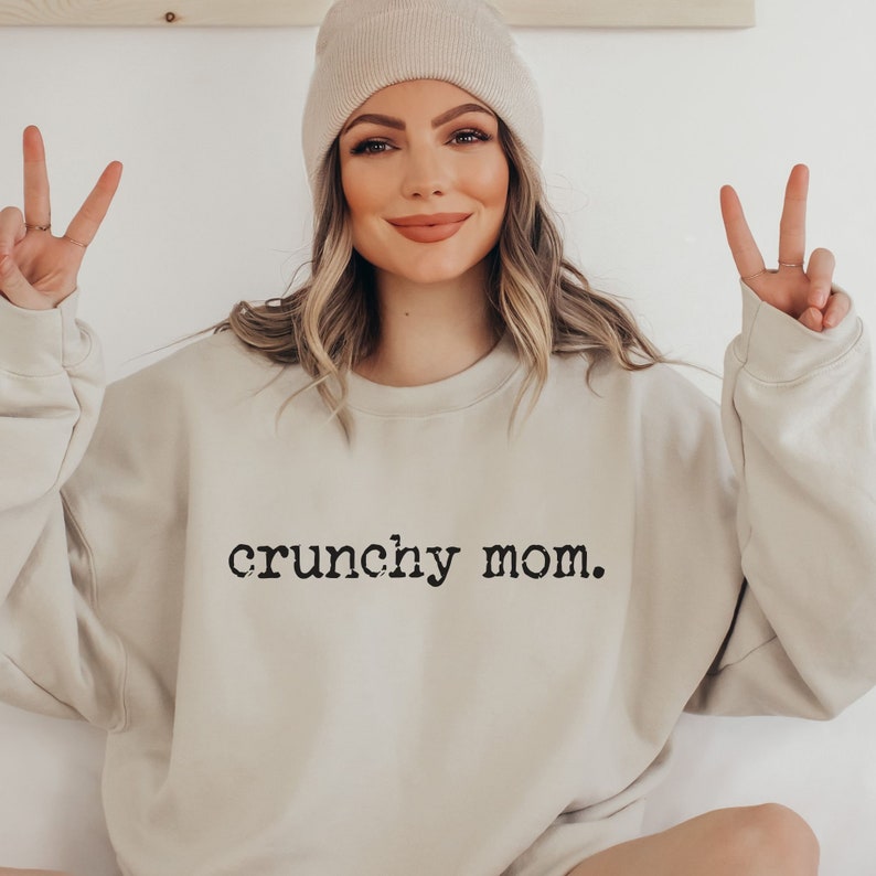 May include: A beige sweatshirt with the text "crunchy mom." printed in black on the front. The model is wearing a beige beanie and is making a peace sign with both hands.