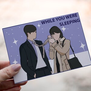 Può includere: Illustrazione di due persone su una cartolina con la scritta "WHILE YOU WERE SLEEPING". Lo sfondo è viola chiaro con stelle bianche. Una persona indossa un abito e una cravatta, e l'altra un cappotto beige.