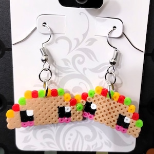 May include: A pair of dangle earrings featuring a taco design made with colorful perler beads. The taco is brown with yellow, red, green, and pink accents. The earrings are attached to silver hooks.