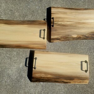 May include: Three wooden serving trays with natural wood grain and black metal handles. The trays are different sizes and have a rustic, handcrafted look.