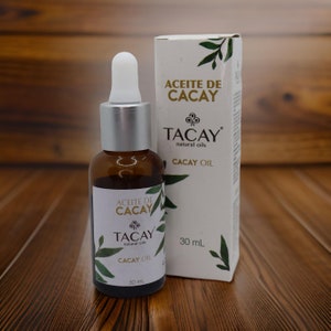 May include: A 30 ml bottle of Cacay oil with a dropper. The bottle is brown glass with a silver cap and a white label. The label has the text "Aceite de Cacay" and "Tacay natural oils" in gold lettering. The bottle is sitting on a wooden surface.