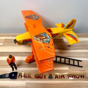 May include: A vintage toy airplane in bright orange and yellow, with colorful decals. The toy includes a small pilot figure, a black ladder, and a black accessory. The words "FALL GUY AIR SHOW" are printed on the surface.