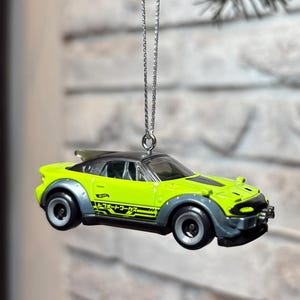 May include: A lime green and dark grey toy sports car ornament hanging from a silver chain. The car features black racing stripes, a rear spoiler, and Japanese text on the side panel. The vehicle has a low-profile design with dark grey wheel arches.