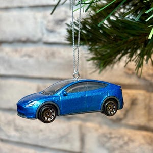 May include: A blue toy car Christmas ornament with a silver hanging chain. The miniature car has black wheels and a clear windshield. The ornament is hanging from a green Christmas tree branch.