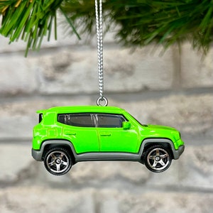 May include: A lime green toy SUV ornament hanging from a silver string. The vehicle features dark tinted windows, grey wheel arches, and silver five-spoke wheels. The body is painted in a bright, glossy neon green finish with a compact, boxy design.