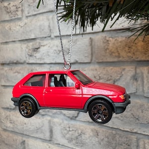 May include: A red toy car Christmas ornament with black wheels and a silver chain. The car has a detailed design, including windows and interior features. The ornament is suspended against a brick wall.