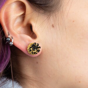May include: A pair of gold stud earrings with black beads. The earrings are worn in the earlobe of a person with purple hair.