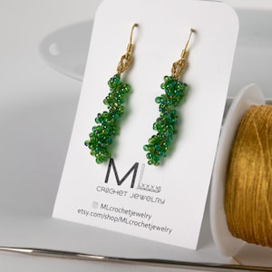 May include: Green beaded earrings with gold hooks. The earrings are crocheted and have a spiral design. The earrings are displayed on a white card with the text "ML Crochet Jewelry" and a website address.