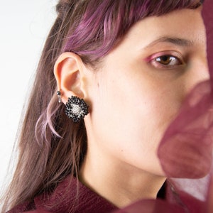 Pode incluir: Um close-up do rosto de uma pessoa com cabelo rosa, usando brincos de contas pretas e prateadas. A pessoa está parcialmente obscurecida por um tecido transparente bordô.
