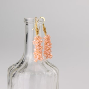 May include: A pair of pink beaded earrings with gold hooks. The earrings are hanging from a clear glass bottle.