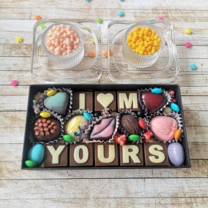 May include: A black box filled with chocolate squares that spell out "I love you". The chocolates are decorated with different colors and designs, including hearts, lips, and flowers. There are two small glass dishes with pink and yellow sprinkles on the table.