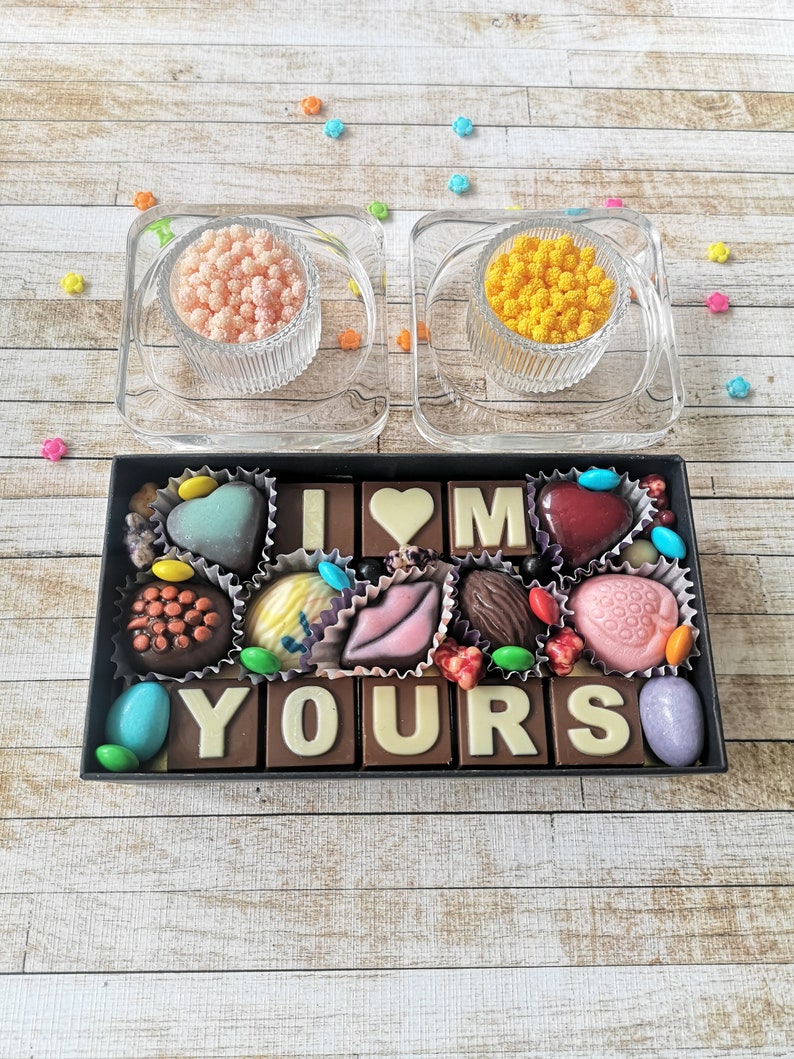 May include: A black box filled with chocolate squares that spell out "I love you". The chocolates are decorated with different colors and designs, including hearts, lips, and flowers. There are two small glass dishes with pink and yellow sprinkles on the table.