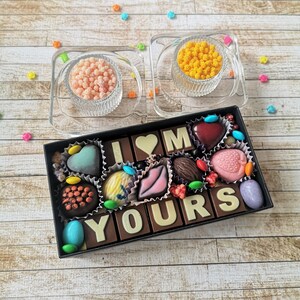 May include: A box of assorted chocolates arranged to spell out "I LOVE YOU" with a heart-shaped chocolate in the center. The box is on a wooden surface with small colorful sprinkles scattered around.
