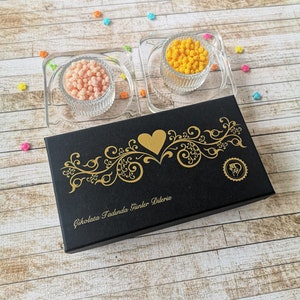 May include: A black box with gold lettering that says "Cikolata Tadinda Gunler Dileriz" and a gold heart design. Two small glass bowls filled with orange and pink sprinkles are on top of the box.
