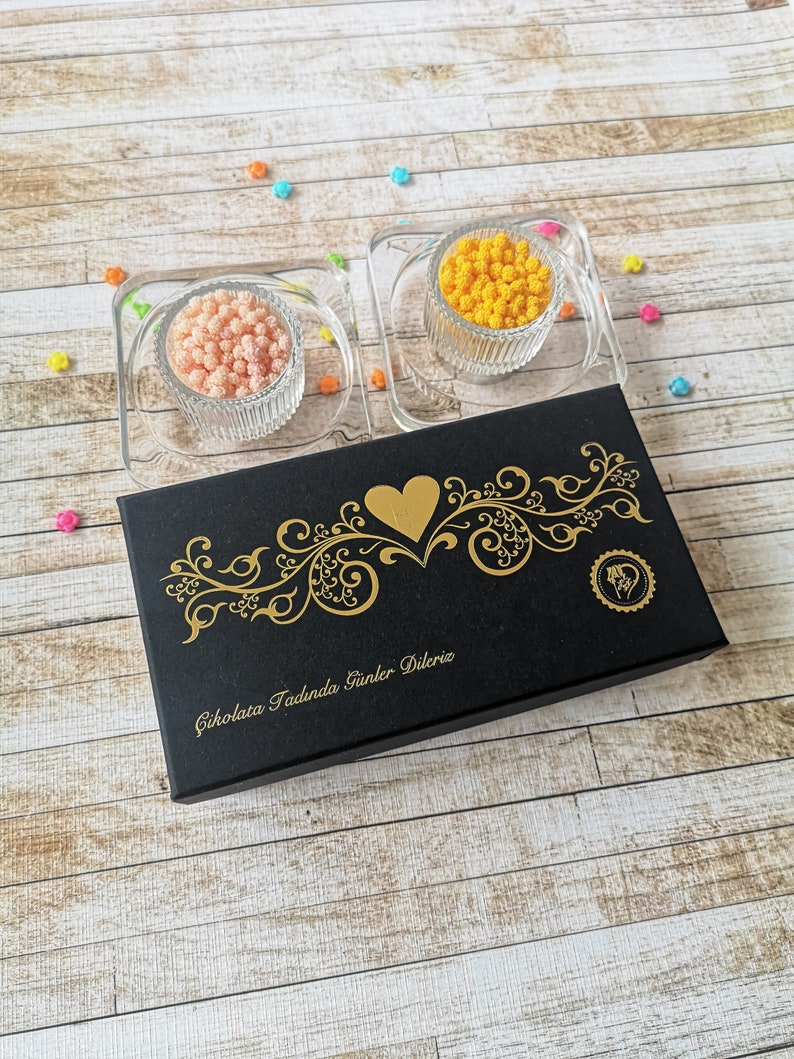 May include: A black box with gold lettering that says "Cikolata Tadinda Gunler Dileriz" and a gold heart design. Two small glass bowls filled with orange and pink sprinkles are on top of the box.