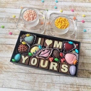 May include: A box of assorted chocolates arranged to spell "I love you". The chocolates are in a variety of shapes and colors, including hearts, lips, and letters. The box is surrounded by small, colorful sprinkles.